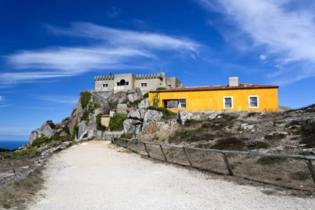 Yellow house and stone building on rocky hill under blue sky with wispy clouds.