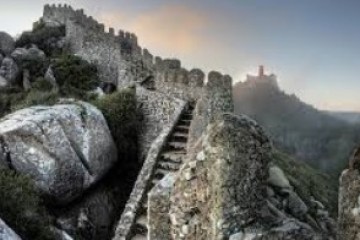 Stone fortress with a stairway, large rocks, and distant view of a red castle on a hill at sunset.