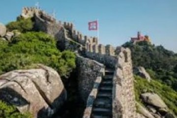 Stone castle wall with staircase, surrounded by greenery, red flag atop, blue sky.