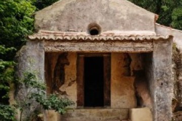 Small stone chapel with stairs and cross, surrounded by lush greenery.