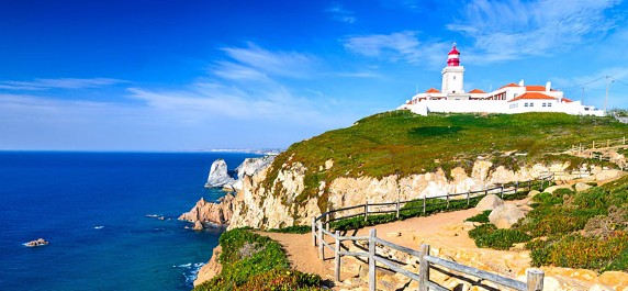 Lighthouse on a cliffside with a wooden fence, overlooking a blue ocean under a clear sky.