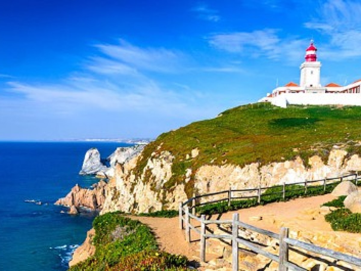 Lighthouse on a cliffside with a wooden fence, overlooking a blue ocean under a clear sky.