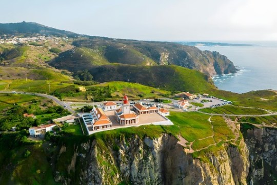 Aerial view of a coastal lighthouse on cliffs with green landscape and ocean.