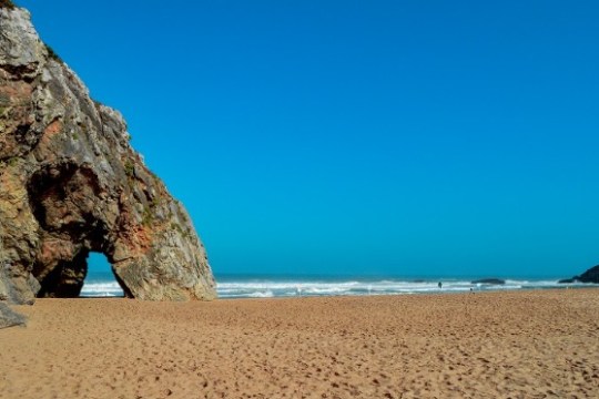 Rock formation with arch on sandy beach, ocean waves, clear blue sky.