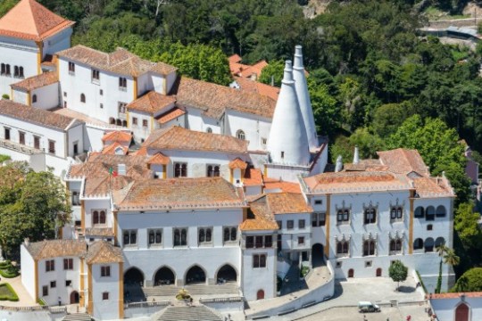 Aerial view of a historic white palace with orange roofs and two large conical chimneys.