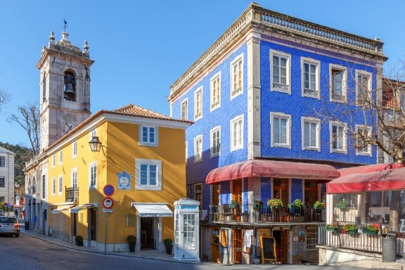 Colorful buildings on a sunny street, blue and yellow facades with a tower in the background.