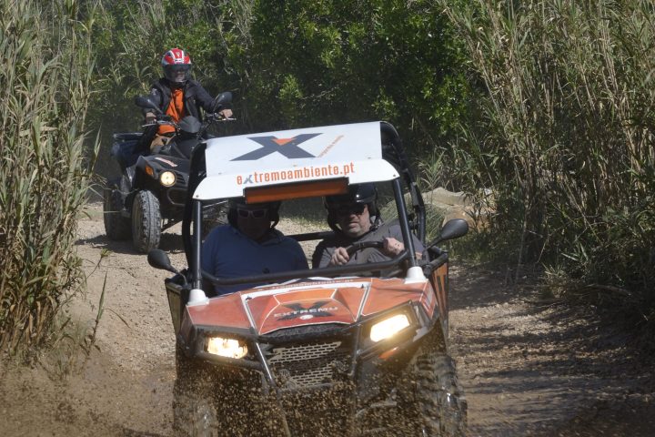 Two off-road buggies driving on a muddy dirt path with greenery around.