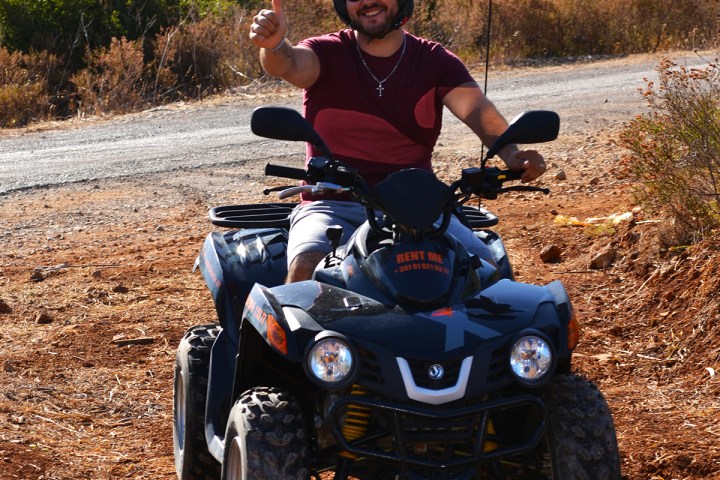 Person on a quad bike giving a thumbs-up on a dirt path surrounded by dry vegetation.