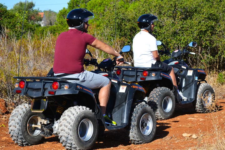 Two people on ATVs wearing helmets, riding on a dirt path surrounded by bushes.