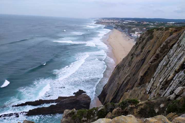 View of ocean waves hitting a sandy beach from a high cliff under a cloudy sky.