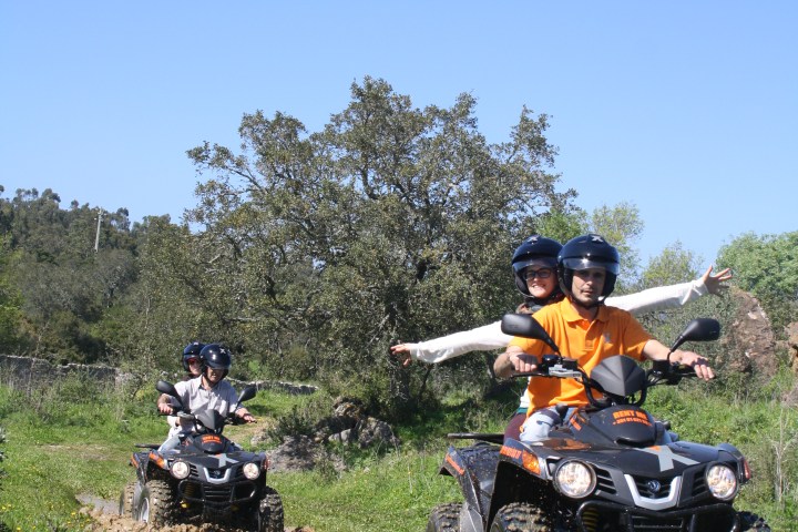 Two people ride ATVs on a grassy trail under a clear blue sky.