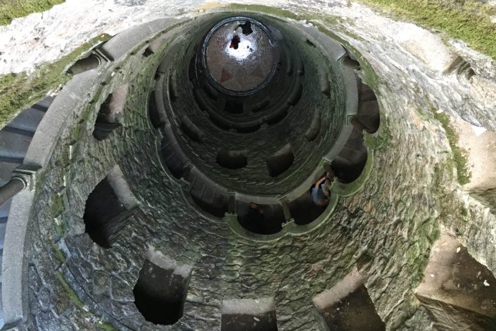 View looking up a stone spiral staircase with mossy walls and circular openings.