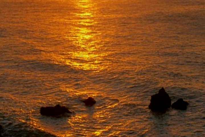 Sunset over the ocean with rocks and waves, casting a golden reflection.