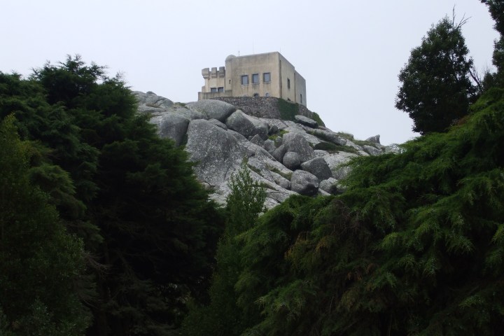 A beige building atop a rocky hill, surrounded by lush trees under a cloudy sky.