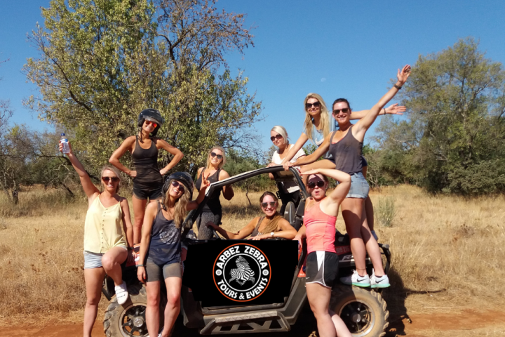 Group of women posing with an ATV in a sunny, grassy area with trees in the background.