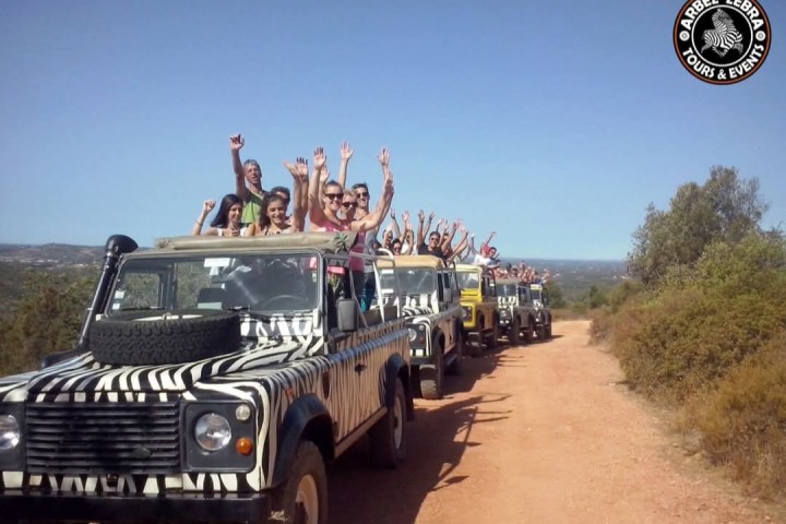 People in zebra-striped jeeps on a dirt road, cheering with raised arms under a clear blue sky.