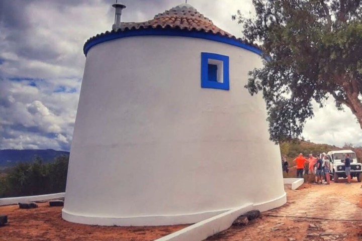 White cylindrical building with a blue window under cloudy skies, surrounded by trees and a parked vehicle.