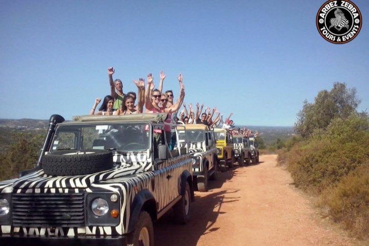 People in open-top safari vehicles with zebra patterns on a dirt road, raising hands.