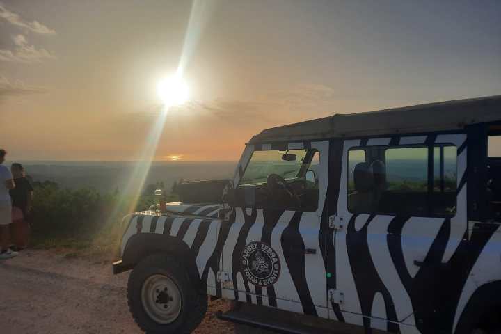 Zebra-striped off-road vehicle parked at sunset with people and scenic landscape visible.
