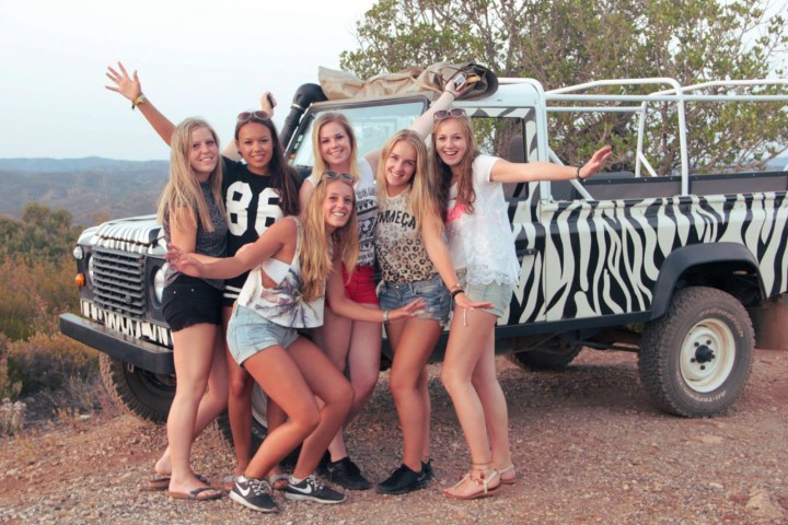 Six smiling women pose in front of a zebra-striped vehicle on a dirt road with hills in the background.