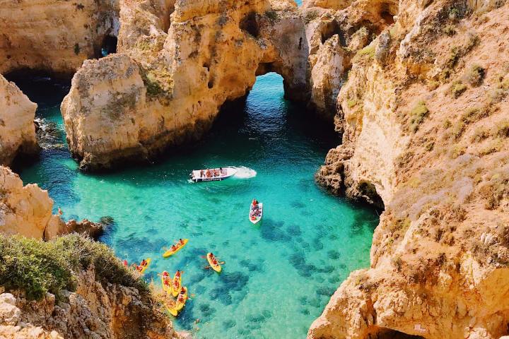 Cliffside cove with turquoise water, kayaks, a boat, and people exploring the rocky formations.
