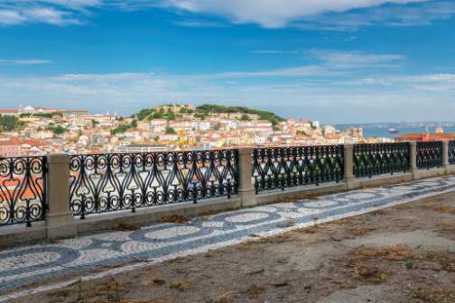 Scenic cityscape viewed from a patterned balcony overlooking a hillside with colorful buildings and a blue sky.