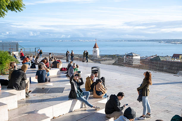 People sitting and standing on a terrace overlooking a body of water and distant cityscape.