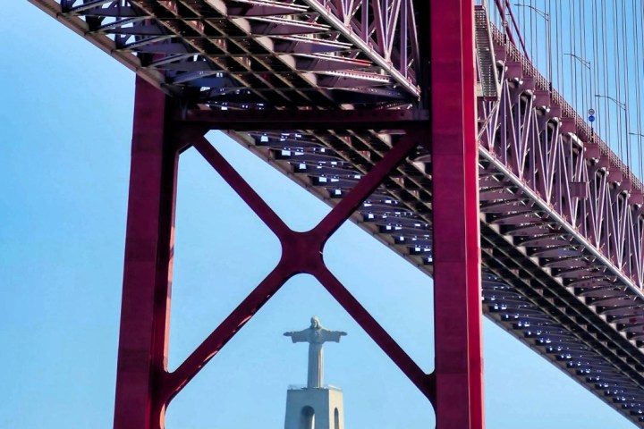 Red bridge framing a statue with outstretched arms against a blue sky.