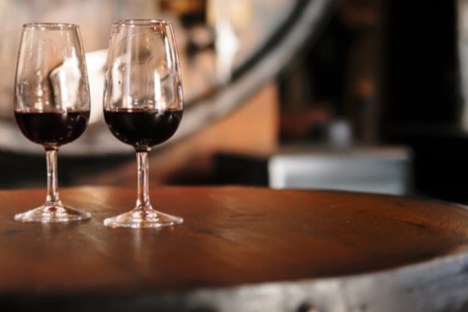 Two glasses of red wine on a wooden barrel in a dimly lit room.