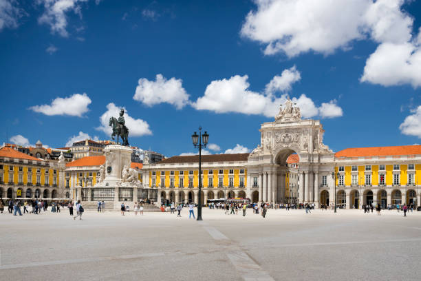 Wide European plaza with statue, yellow buildings, and archway under a blue sky with clouds.