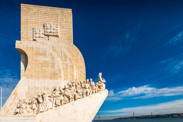 Stone monument with figures overlooking water under a blue sky.