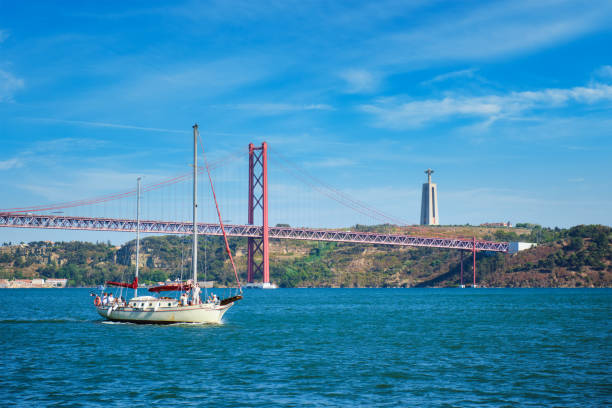 Sailboat on river with red suspension bridge and large statue in the background against a blue sky.