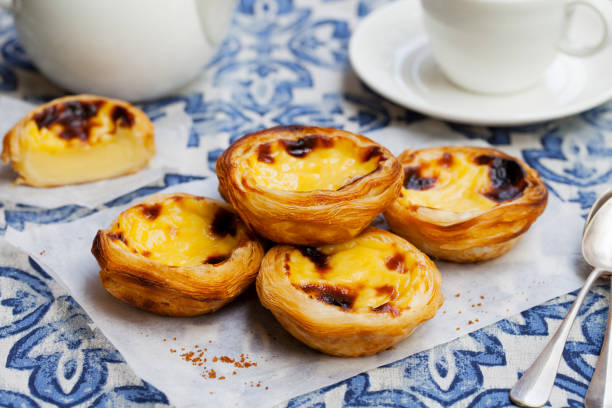 Four Portuguese custard tarts on parchment with a teacup and teapot on a blue patterned cloth.