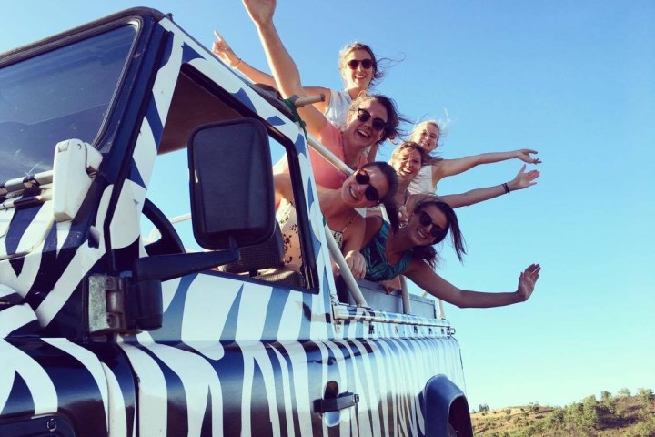 Group of people smiling, waving out of zebra-striped vehicle under a clear blue sky.