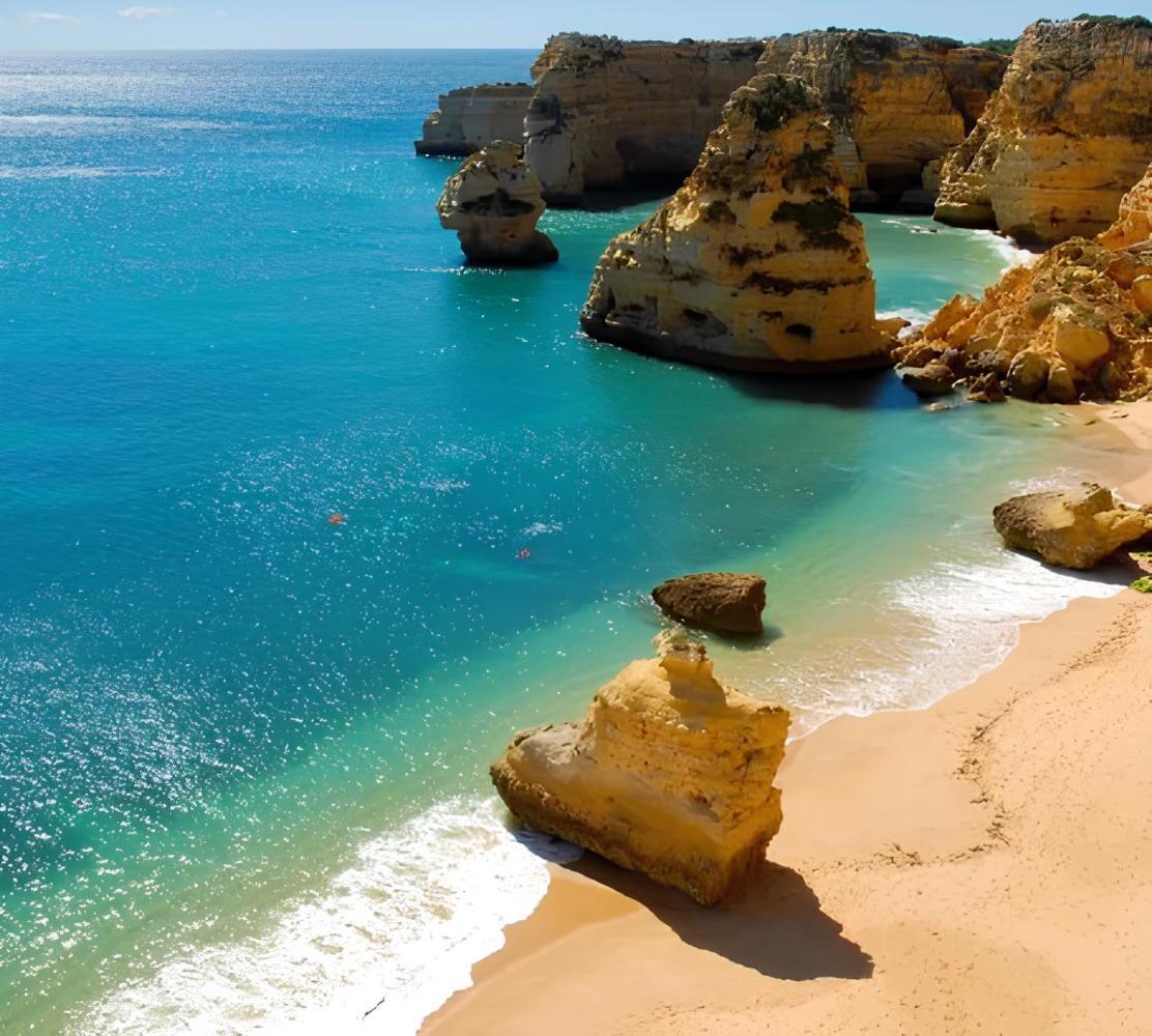 Sunny beach with turquoise water and large rock formations along the shore.