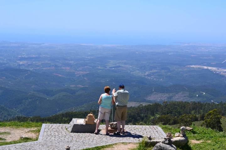 Two people admire a vast mountain view with clear skies and ocean beyond.