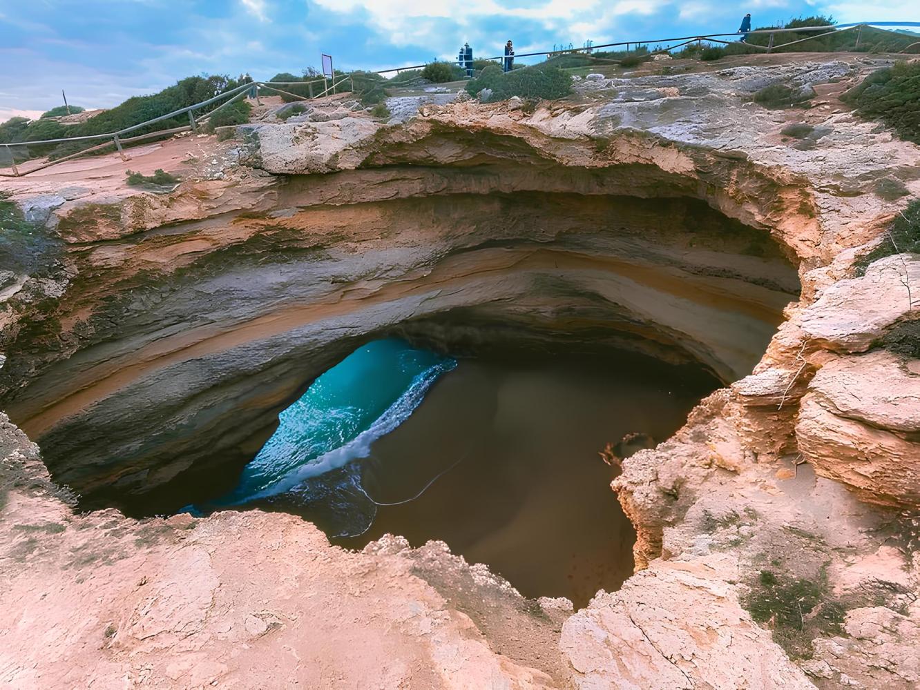 Natural coastal cave with water and people on top, cloudy sky.