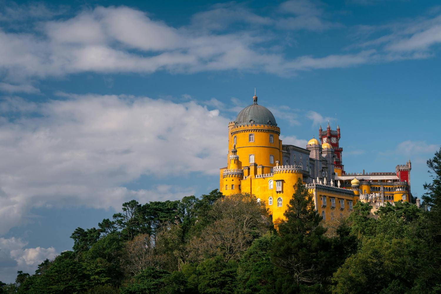 Colorful castle with yellow facade on a hill surrounded by trees, under a partly cloudy blue sky.
