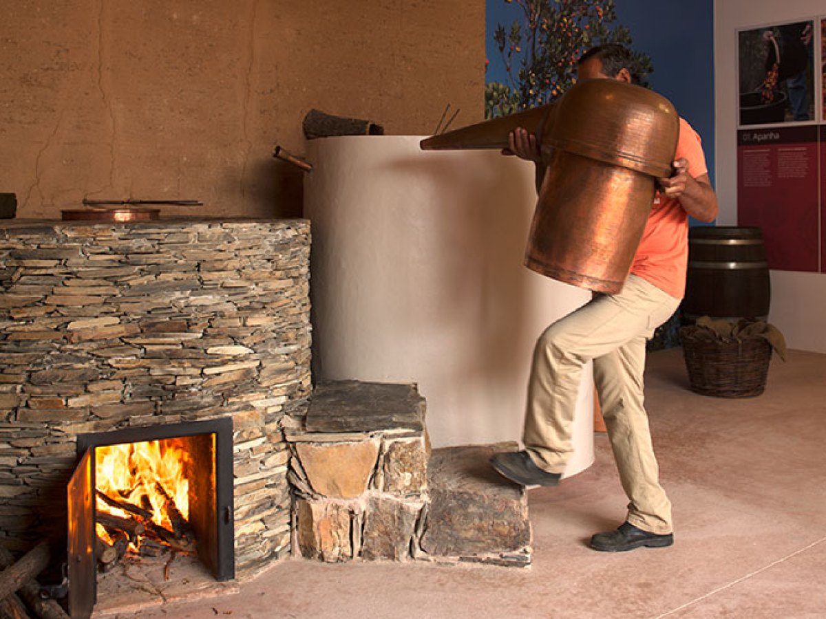 Person carrying a large copper object near a stone oven with fire inside.